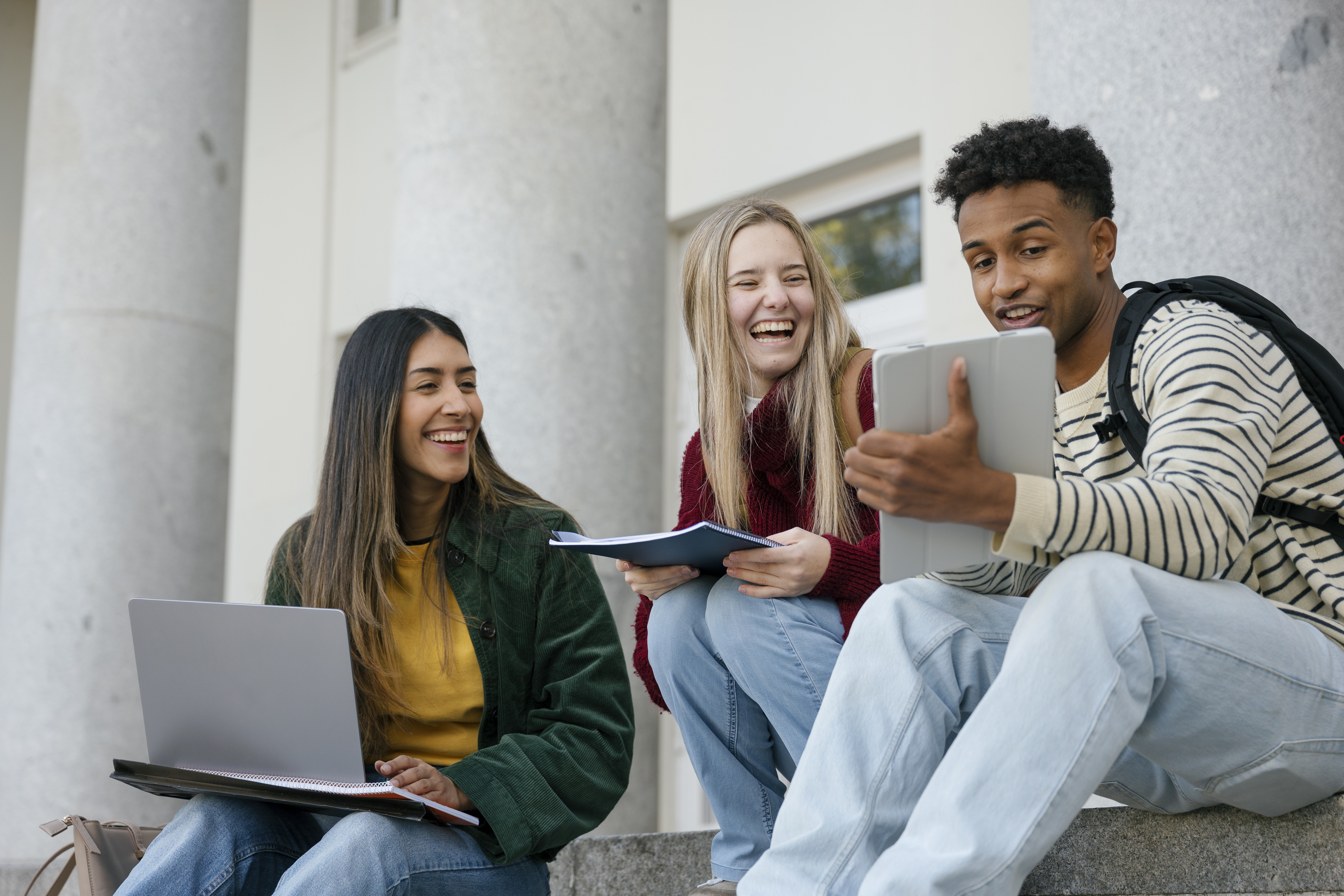 Three diverse university students sitting on campus stairs, talking and smiling while looking at a digital tablet and laptop, collaborating in an urban environment